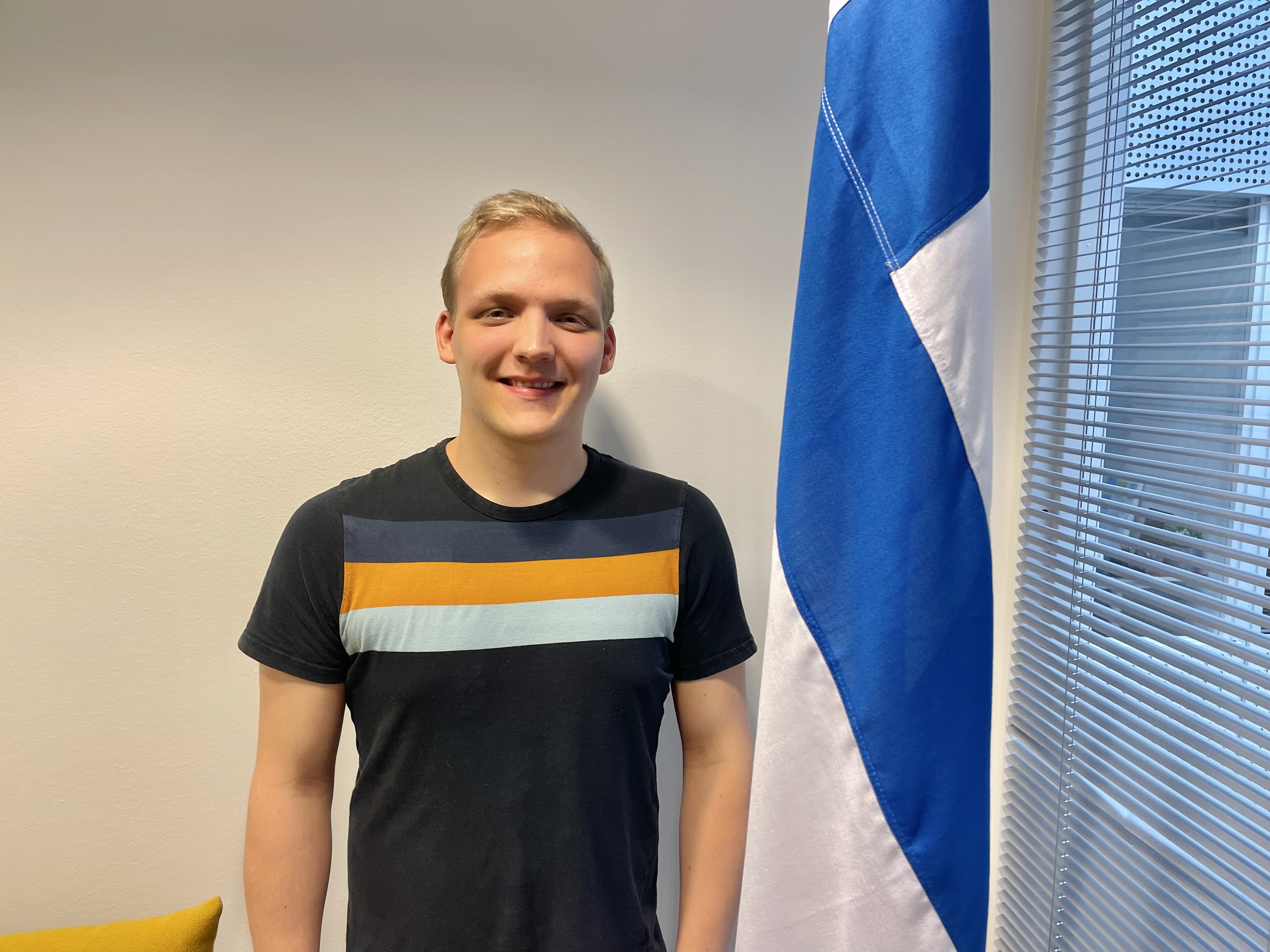 Musiikin opiskelija Peetu Karjalainen hymyilee Suomen lipun vieressä.