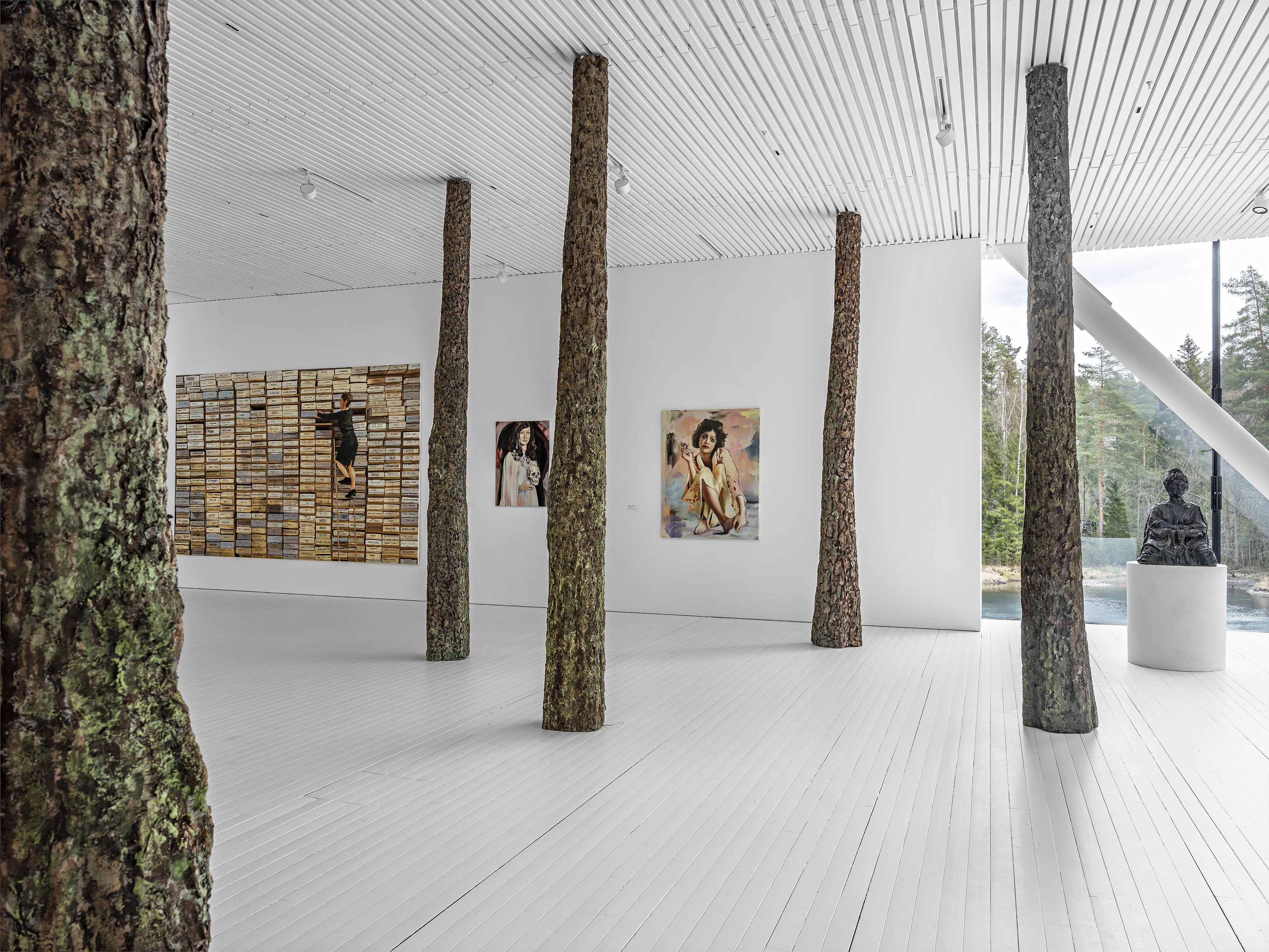 Taidenäyttelyn huoneessa kolme taulua seinällä. Salissa viisi puunrunkoa lattiasta kattoon.
