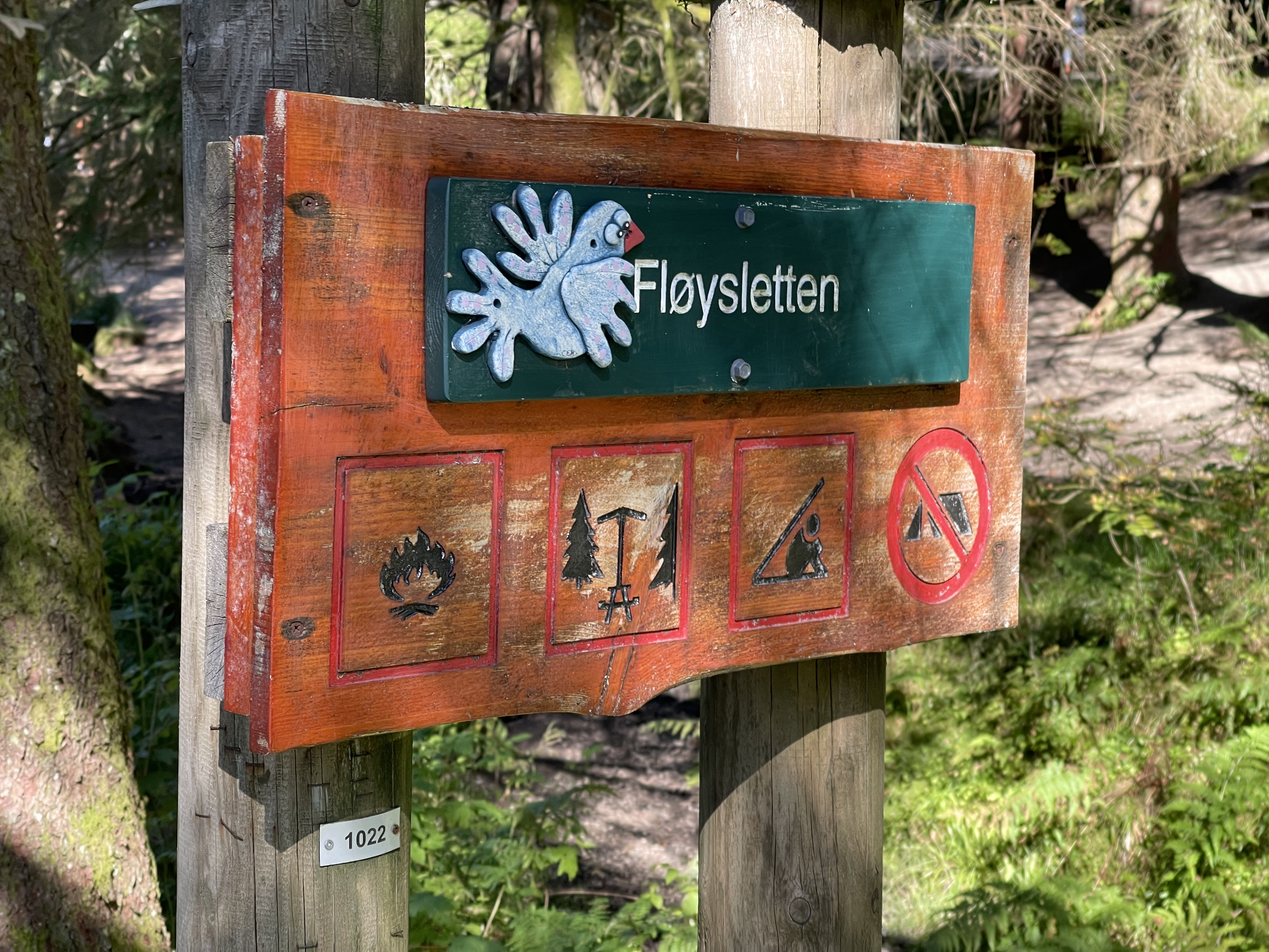Metsän keskellä kahteen paksuun puutolppaan kiinnitetty kyltti. Kyltin yläosassa näkyy valkoinen kyyhkynen ja paikannimi Fløysletten. Alaosassa neljä kuvaketta: nuotiopaikka, taukopöytä, laavupaikka ja telttailukielto.